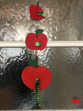 Selbstgebastelte Girlande mit drei roten Papieräpfeln, die übereinander an einer Schnur hängen. Jeder Apfel hat einen aus grünen Papierstreifen gefalten Wurm.